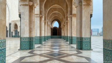 Long pointed-arch passage with stone piers, blue-green zellij bases, patterned floor, and distant dark doorway.