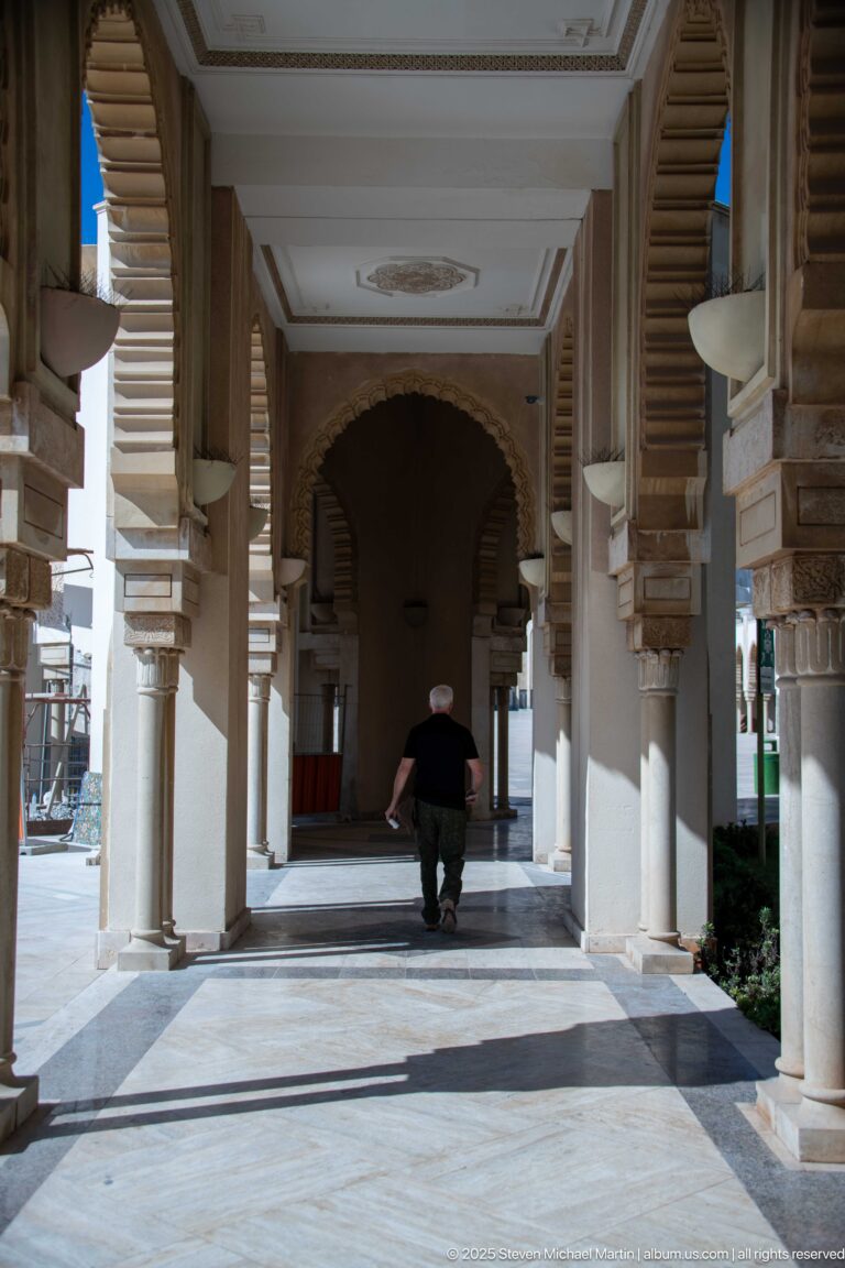 Shaded mosque arcade with repeated columns and brackets, with one visitor walking toward a dark arch.