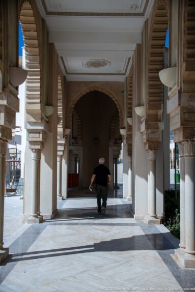 Shaded mosque arcade with repeated columns and brackets, with one visitor walking toward a dark arch.