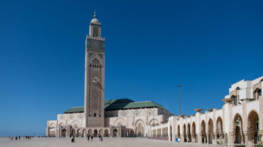 Full Hassan II Mosque with tall minaret, green-roofed main building, arcade, plaza, and small visitors.