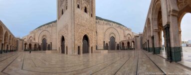 Courtyard with pale mosque walls, tall arched doors, minaret base, surrounding arcades, and patterned paving.
