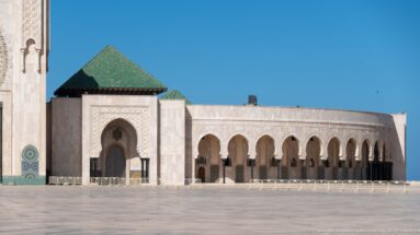 Green-roofed mosque pavilion beside a long arcade across an open paved courtyard.