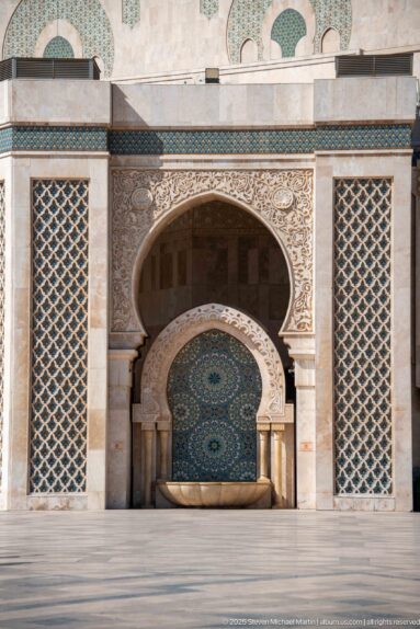 Broad courtyard fountain with semicircular mosaic panel, long stone basin, carved arch, and lattice screens.