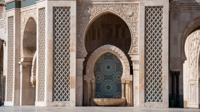 Blue-green wall fountain beneath a carved horseshoe arch, flanked by tall patterned screens.