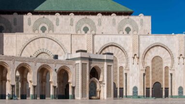 Broad mosque courtyard with pale façade, repeated arches, covered arcade, portals, and green roof.