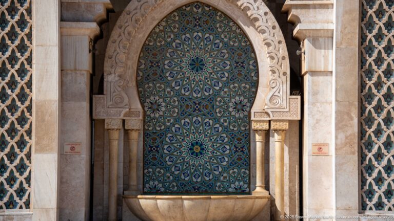 Arched blue-green mosaic fountain niche with geometric rosettes above a shallow stone basin.