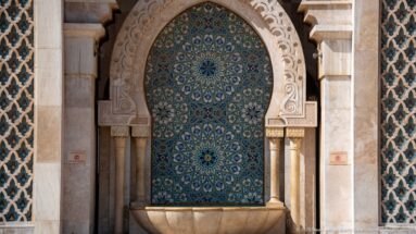 Arched blue-green mosaic fountain niche with geometric rosettes above a shallow stone basin.