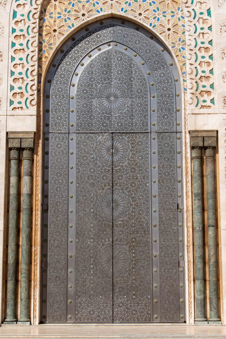 Large arched dark metal door covered with circular geometric patterns and framed by mosaic decoration.