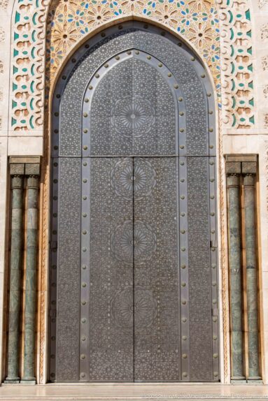 Large arched dark metal door covered with circular geometric patterns and framed by mosaic decoration.