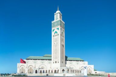 Frontal Hassan II Mosque exterior with tall minaret, green roofs, Moroccan flag, and clear blue sky.