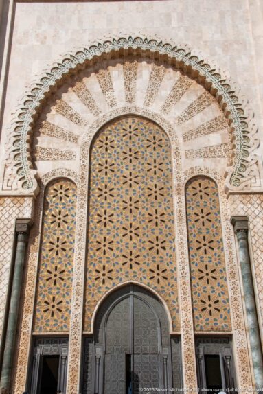 Large decorated mosque entrance with dark metal door, patterned panels, scalloped arch, and side columns.