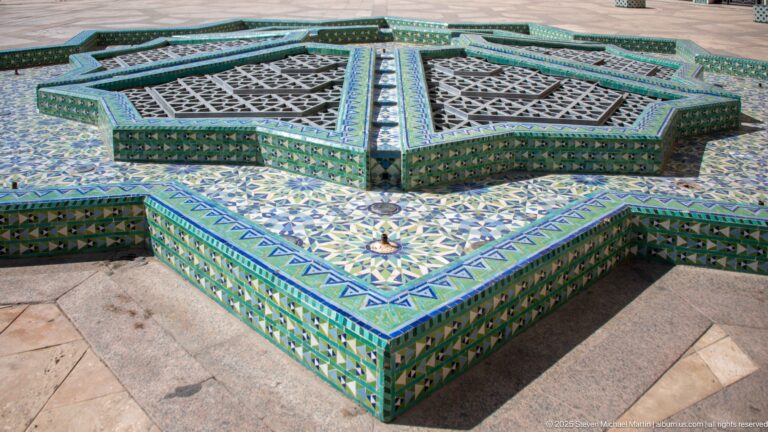 Star-shaped tiled courtyard fountain with patterned sides, metal grilles, and small central nozzle.