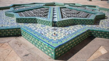 Star-shaped tiled courtyard fountain with patterned sides, metal grilles, and small central nozzle.