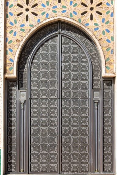 Arched dark metal door with geometric lattice and colorful mosaic ornament above.