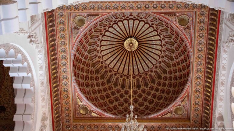 Square decorated ceiling panel with radial center, geometric borders, and part of a chandelier.