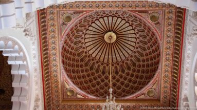 Square decorated ceiling panel with radial center, geometric borders, and part of a chandelier.