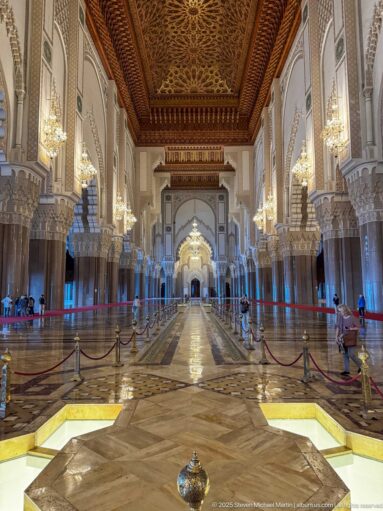 Broad mosque interior with patterned floor, wooden ceiling, chandeliers, and receding columns.