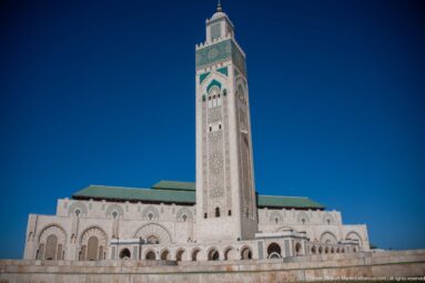 Frontal low-angle view of the Hassan II Mosque minaret above the main arched façade and green roofs.