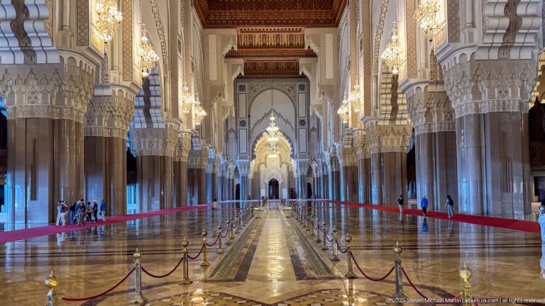 Symmetrical central aisle with stone columns, chandeliers, polished floor, and red barriers.