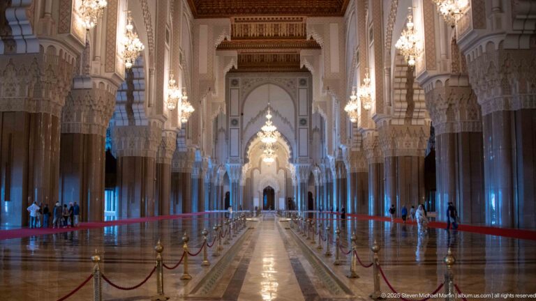 Long mosque aisle with repeating columns, scalloped arches, chandeliers, and a bright distant arch.