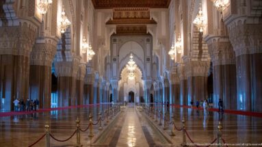 Long mosque aisle with repeating columns, scalloped arches, chandeliers, and a bright distant arch.