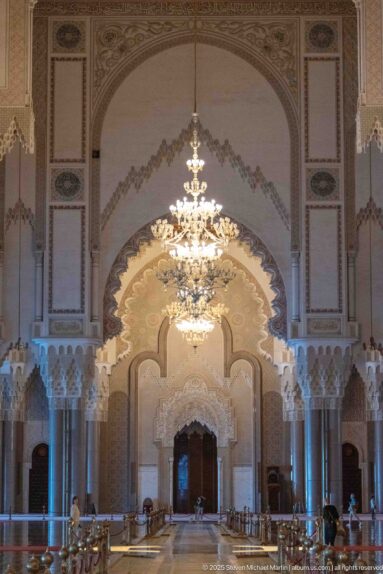 Axial mosque interior with chandeliers, repeated arches, polished floor, and visitors.