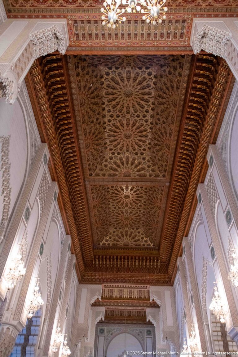 Large carved wooden ceiling with dense interlaced floral and geometric relief, deep border bands, and chandelier at the edge.