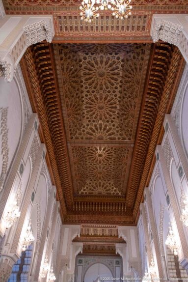 Large carved wooden ceiling with dense interlaced floral and geometric relief, deep border bands, and chandelier at the edge.