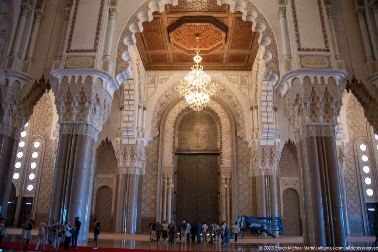 Tall dark interior portal framed by massive columns and scalloped arches, with chandelier and visitors below.
