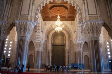 Tall dark interior portal framed by massive columns and scalloped arches, with chandelier and visitors below.