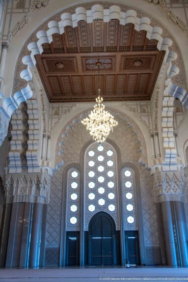 Monumental dark doorway framed by scalloped arches, carved capitals, tall columns, chandelier, and coffered wooden ceiling.