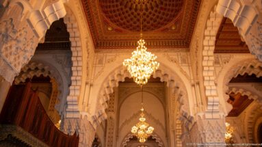 Warm-lit sequence of scalloped arches, carved capitals, chandeliers, and decorated ceilings inside the prayer hall.