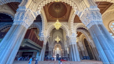 Low-angle prayer hall view with towering columns, scalloped arches, chandeliers, wood ceilings, and visitors for scale.