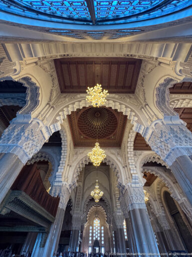 Wide prayer hall interior with massive columns, scalloped arches, carved capitals, chandeliers, and dark wooden ceiling panels.