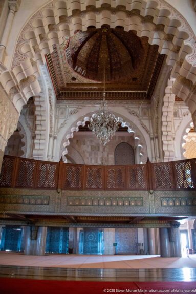 Carved wooden balcony and railing beneath a decorated ceiling bay, chandelier, and scalloped arches inside the mosque.