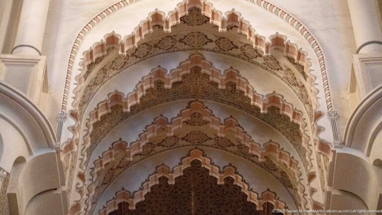 Upward close view of nested scalloped arches with carved ornament and darker woodwork beyond.