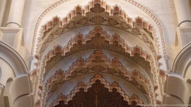 Upward close view of nested scalloped arches with carved ornament and darker woodwork beyond.
