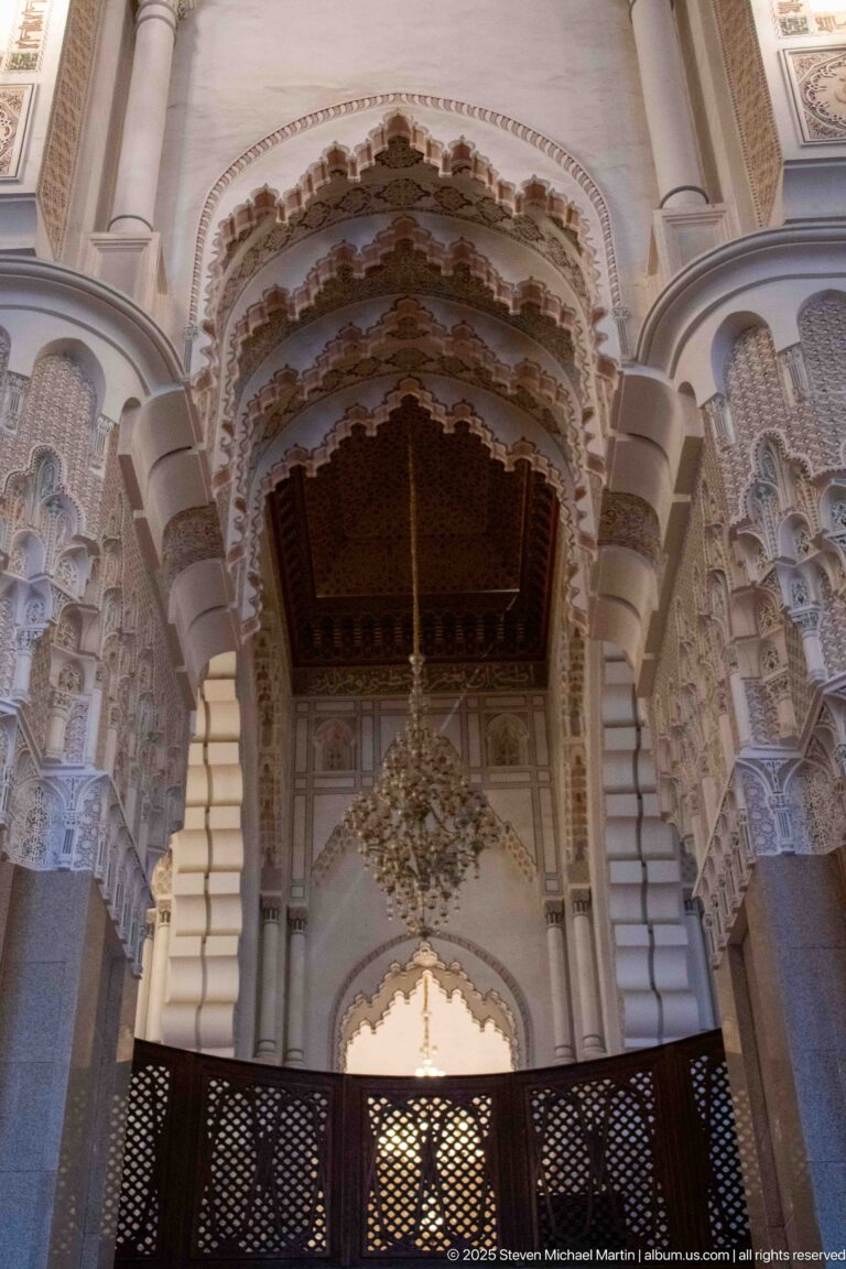 Ornamented scalloped arch with chandelier and carved supports above a dark lattice screen inside the Hassan II Mosque.