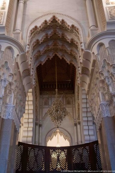 Ornamented scalloped arch with chandelier and carved supports above a dark lattice screen inside the Hassan II Mosque.