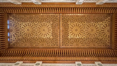 Carved wooden ceiling panel with two large radial rosettes, dense geometric relief, and deep projecting cornice bands.