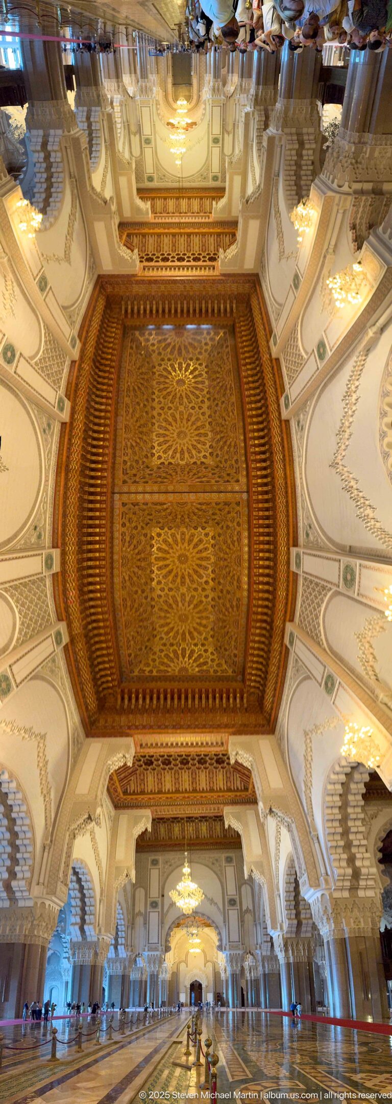 Wide upward view through repeated decorated ceiling bays, arches, chandeliers, and columns in the prayer hall.