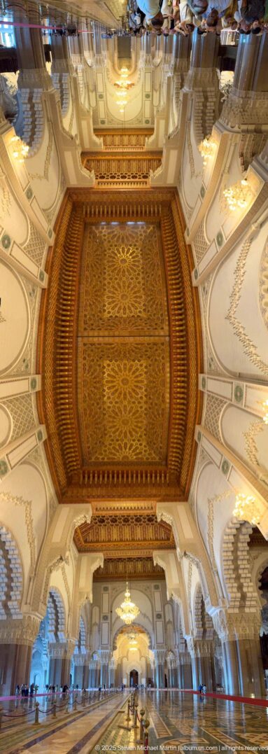 Wide upward view through repeated decorated ceiling bays, arches, chandeliers, and columns in the prayer hall.