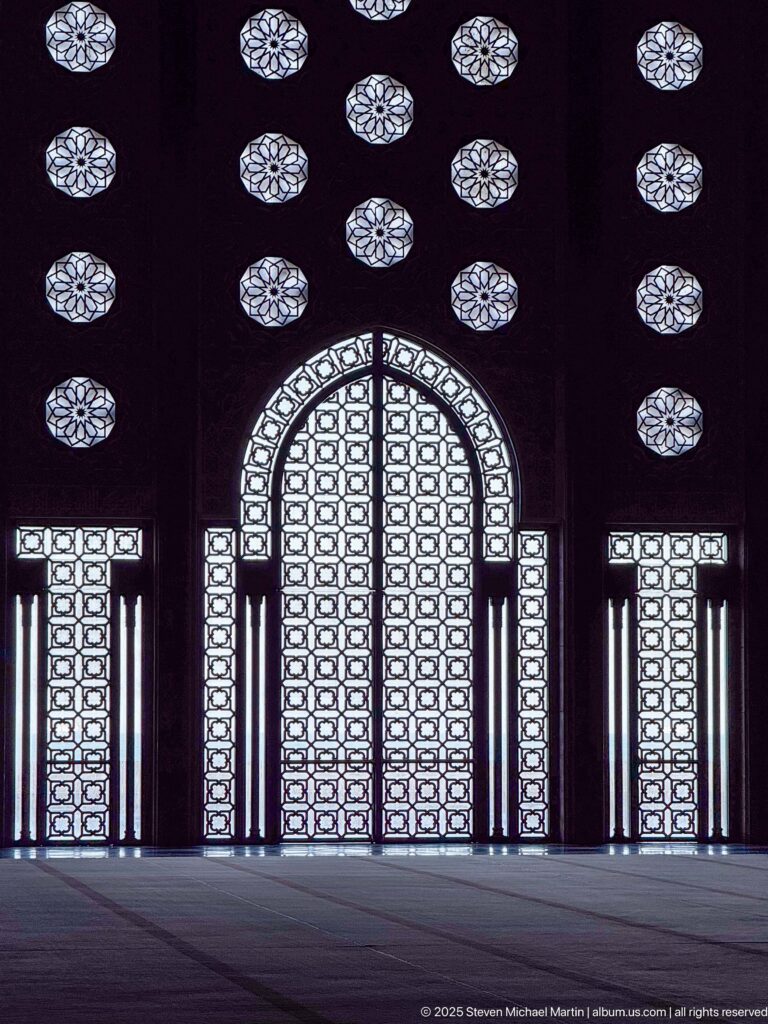 Backlit pointed doorway with patterned screens, flanking rectangular openings, and rows of circular rosettes inside the mosque.