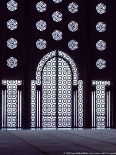 Backlit pointed doorway with patterned screens, flanking rectangular openings, and rows of circular rosettes inside the mosque.