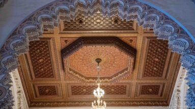 Octagonal decorated ceiling panel with chandelier, carved coffers, and scalloped arch framing inside the mosque.