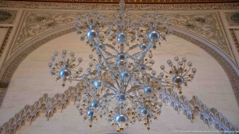 Large branching chandelier hangs in front of a richly decorated arch with scalloped and geometric ornament.