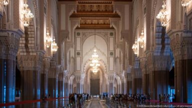Long central view through the Hassan II Mosque prayer hall with chandeliers, arches, columns, visitors, and distant focal arch.
