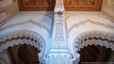 Carved interior pier between scalloped arches beneath a richly decorated wooden ceiling in the Hassan II Mosque.