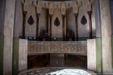 Curved gallery with carved balustrade, stone columns, green tilework, and shadowed lower passage inside the mosque complex.