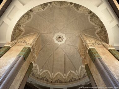 Symmetrical upward view of patterned mosque vault between two monumental stone columns with green decorative bands.